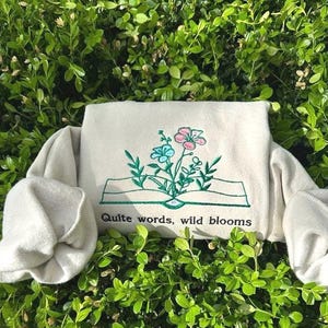 May include: A cream-colored sweatshirt with an embroidered design of a book with flowers and the text "Quite words, wild blooms".