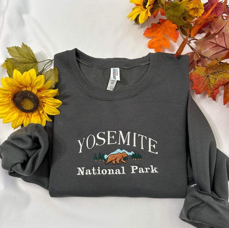 May include: A dark gray sweatshirt with the text "Yosemite National Park" embroidered on the front. The design includes a brown bear walking through a forest scene.