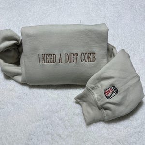 May include: A light grey sweatshirt with the text "I NEED A DIET COKE" embroidered on the front. The sweatshirt has a small embroidered image of a can of Diet Coke on the sleeve.
