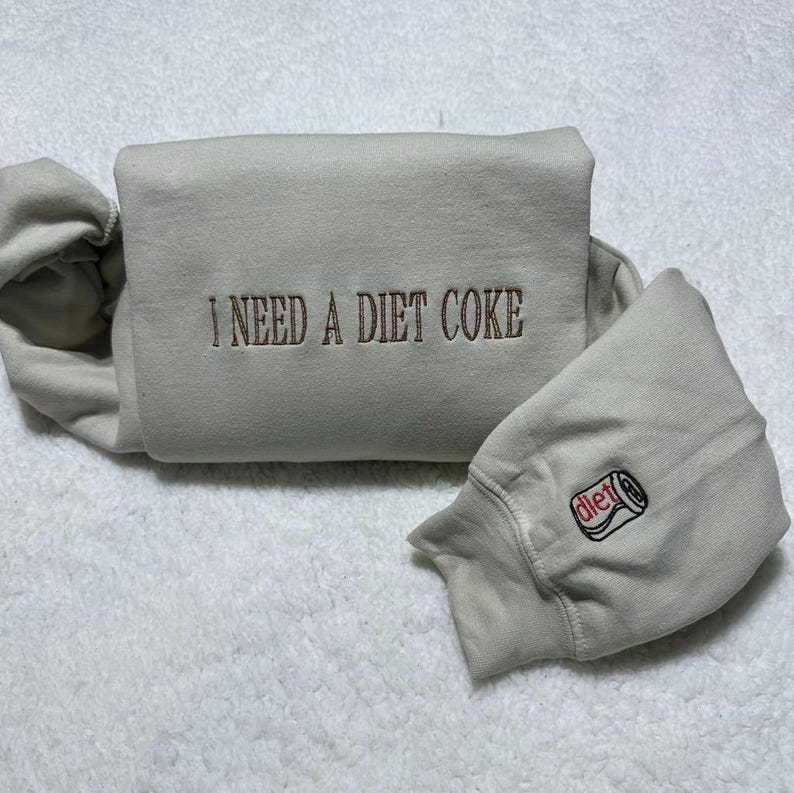 May include: A light grey sweatshirt with the text "I NEED A DIET COKE" embroidered on the front. The sweatshirt has a small embroidered image of a can of Diet Coke on the sleeve.