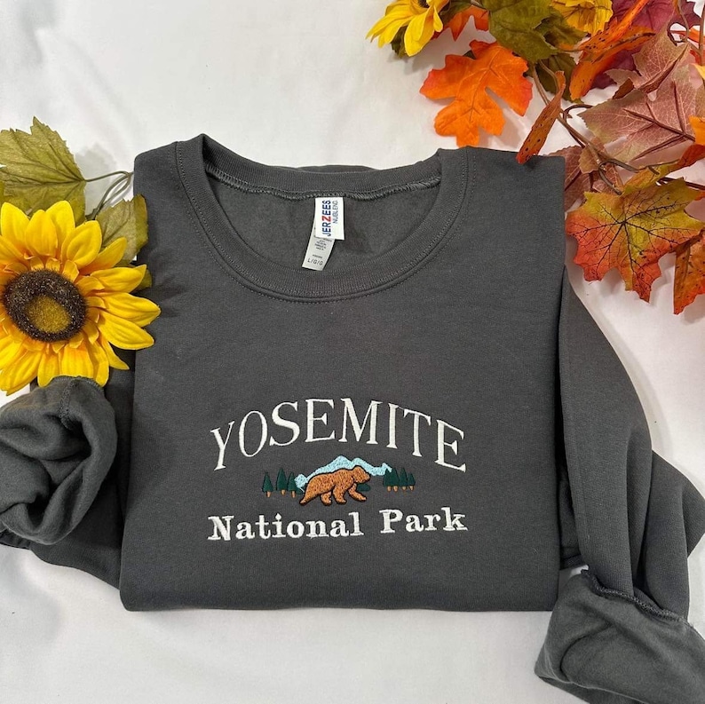 May include: A dark gray sweatshirt with the text "Yosemite National Park" in white embroidery. A brown bear with green trees and blue mountains are embroidered below the text.
