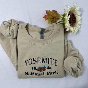 May include: A beige sweatshirt with the text "Yosemite National Park" and an embroidered image of a bear in a forest setting.