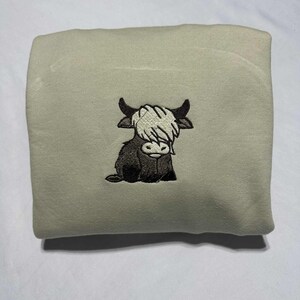 May include: A light green sweatshirt featuring an embroidered Highland cow design. The cow is depicted in black and white, with a shaggy white fringe and black horns. The sweatshirt is folded and presented on a white surface.