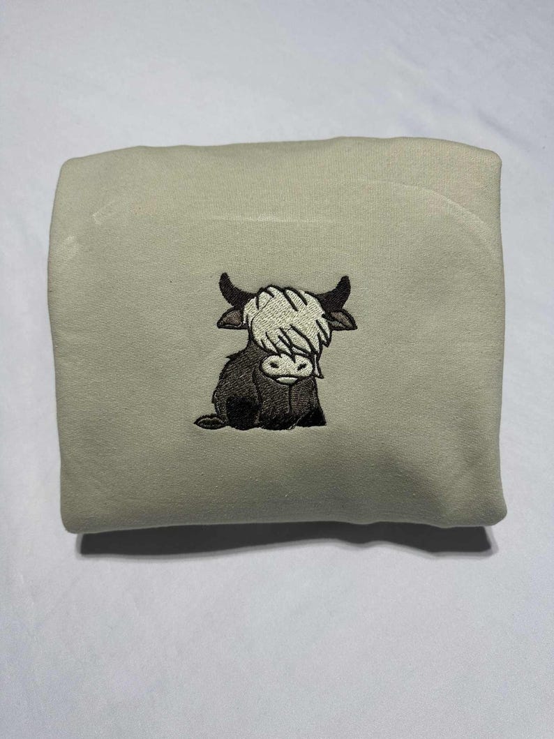 May include: A light green sweatshirt featuring an embroidered Highland cow design. The cow is depicted in black and white, with a shaggy white fringe and black horns. The sweatshirt is folded and presented on a white surface.