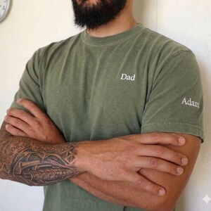 May include: Olive green t-shirt with the word "Dad" embroidered on the chest and "Adam" on the sleeve. The shirt is a solid color and has a classic crew neck design. The person's arms are crossed.