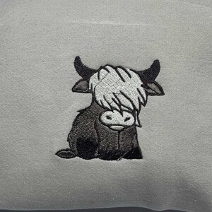 May include: A light gray sweatshirt featuring an embroidered image of a Highland cow. The cow is depicted in shades of black, white, and gray, with a shaggy coat and curved horns. The design is centered on the front of the sweatshirt.
