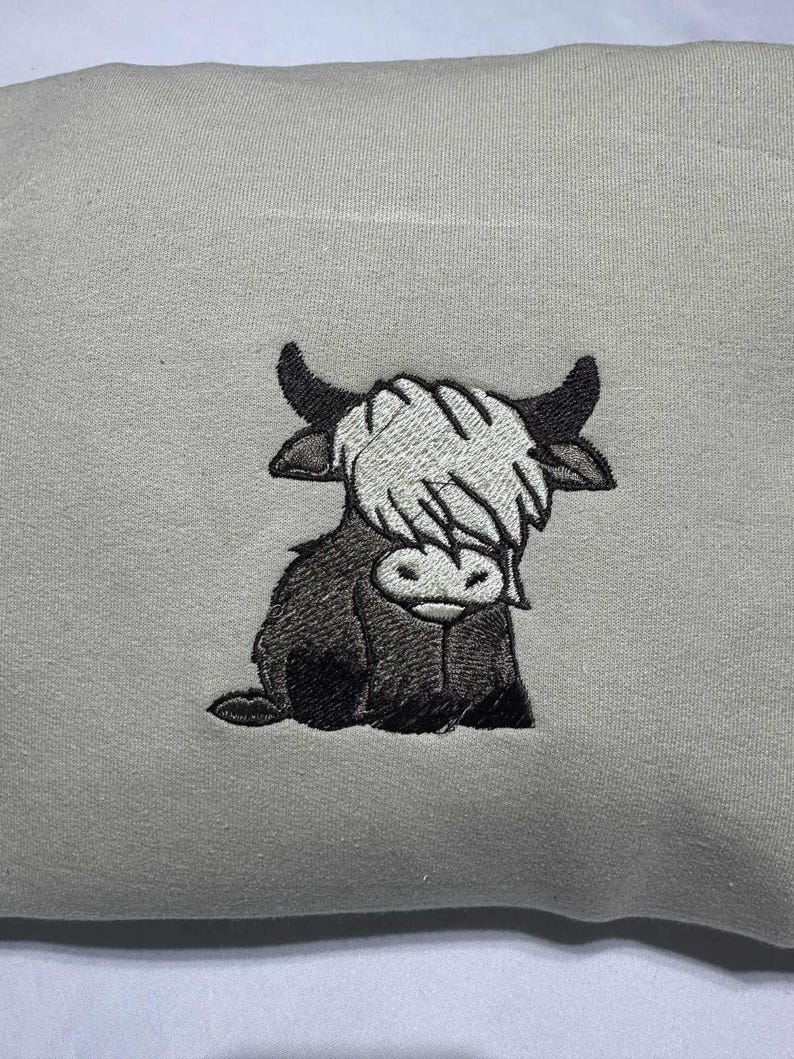 May include: A light gray sweatshirt featuring an embroidered image of a Highland cow. The cow is depicted in shades of black, white, and gray, with a shaggy coat and curved horns. The design is centered on the front of the sweatshirt.