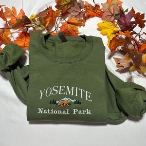 May include: A green sweatshirt with the text "Yosemite National Park" embroidered on the front. The embroidery features a brown bear walking through a forest scene.