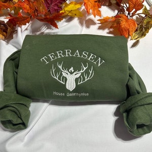 Puede incluir: Una sudadera verde con un logotipo bordado en blanco que dice "TERRASEN" y "House Galathynius" con una cabeza de ciervo estilizada en el centro.