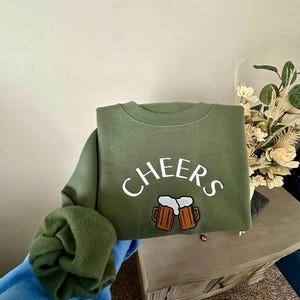 May include: A green sweatshirt with the word "CHEERS" in white lettering and a graphic of two beer mugs.