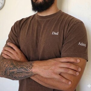 May include: Brown t-shirt with the word "Dad" embroidered on the chest and "Adam" on the sleeve. The shirt is a solid color and has a crew neck. The person wearing the shirt has a tattoo on their arm.