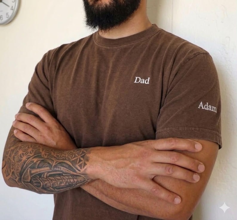 May include: Brown t-shirt with the word "Dad" embroidered on the chest and "Adam" on the sleeve. The shirt is a solid color and has a crew neck. The person wearing the shirt has a tattoo on their arm.
