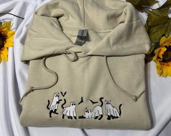Puede incluir: Una sudadera con capucha beige claro con un diseño bordado de cinco gatos blancos vestidos de fantasmas con murciélagos volando a su alrededor.