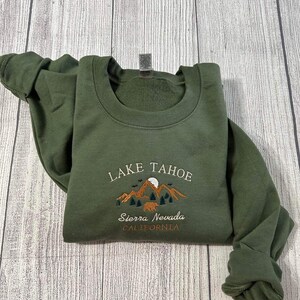 May include: A green sweatshirt with an embroidered design featuring the words "Lake Tahoe", "Sierra Nevada", and "California". The design also includes a mountain range, a moon, and a bear.