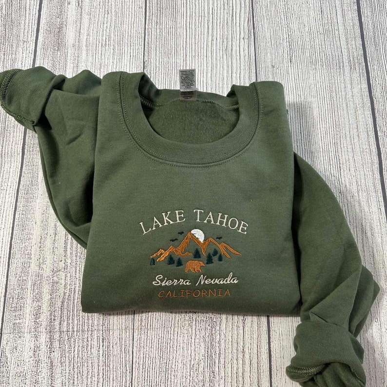 May include: A green sweatshirt with an embroidered design featuring the words "Lake Tahoe", "Sierra Nevada", and "California". The design also includes a mountain range, a moon, and a bear.