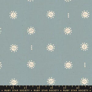 May include: A dusty blue fabric featuring a repeating pattern of cream-colored suns with faces and small triangular motifs. A black ruler at the bottom edge shows measurements in inches, with the text Ruby Star Society printed repeatedly.