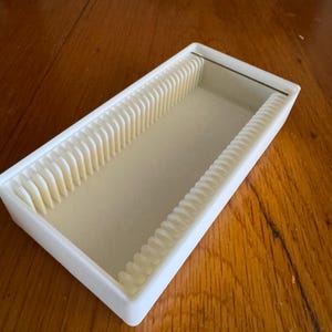 May include: A white plastic slide storage box with vertical dividers. The rectangular box has a light beige interior, designed for microscope slides. It rests on a wooden surface. The box is approximately 15 cm long.