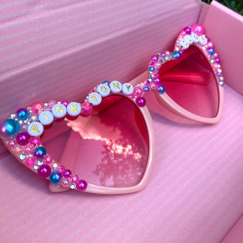 May include: Pink heart-shaped sunglasses with pink lenses and a beaded frame. The frame is decorated with white beads that spell out "PINK PONY CLUB".