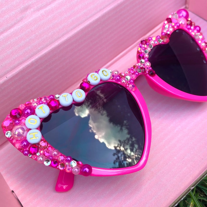 May include: Pink heart-shaped sunglasses with rhinestones and the words "HOT TO GO" spelled out in gold letters on the frame.