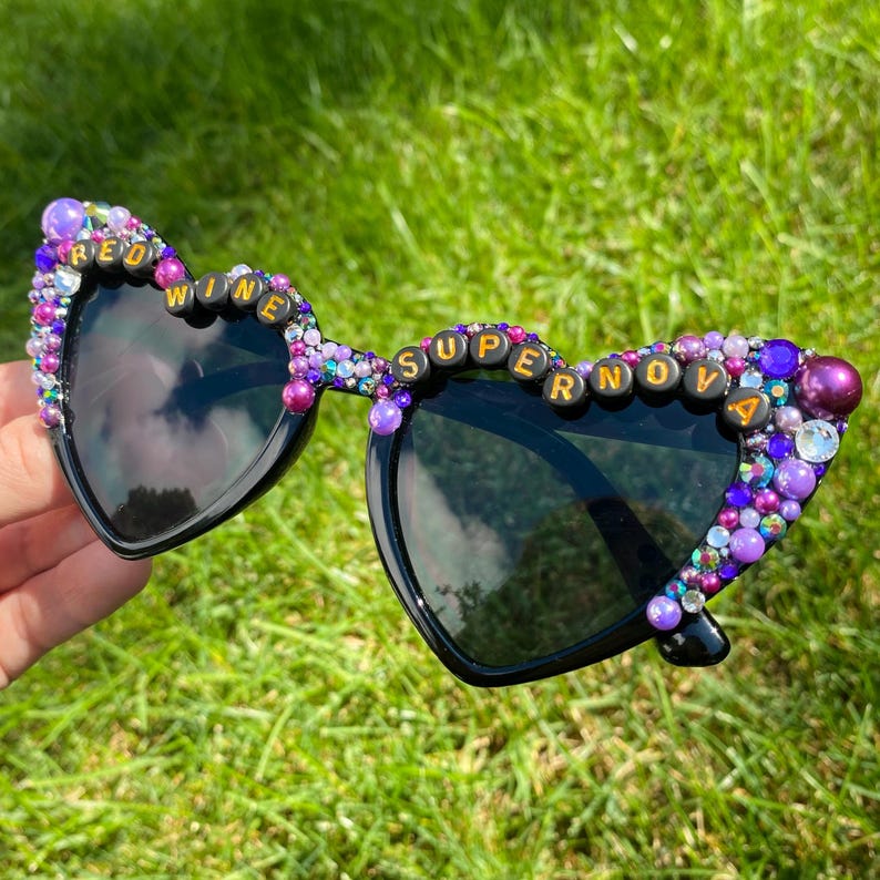 May include: Heart-shaped sunglasses with black frames and purple and pink rhinestones. The rhinestones spell out "Red Wine Supernova".