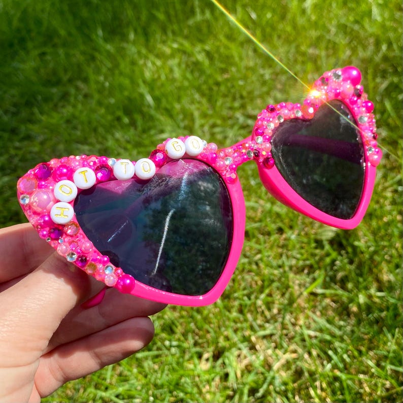 May include: Pink heart-shaped sunglasses with rhinestones and white beads spelling out "HOT TO GO" in gold lettering.