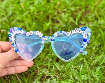 Puede incluir: Gafas de sol en forma de corazón con montura y lentes azul claro. Los bordes están decorados con cuentas azules y blancas y las palabras "LOVE ME TO HEAVEN". Las gafas de sol están sobre un fondo de hierba verde.