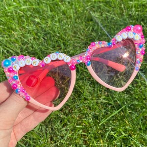 May include: Pink heart-shaped sunglasses with pink, blue, and white beads and the words "Pink Pony Club" spelled out in gold letters.
