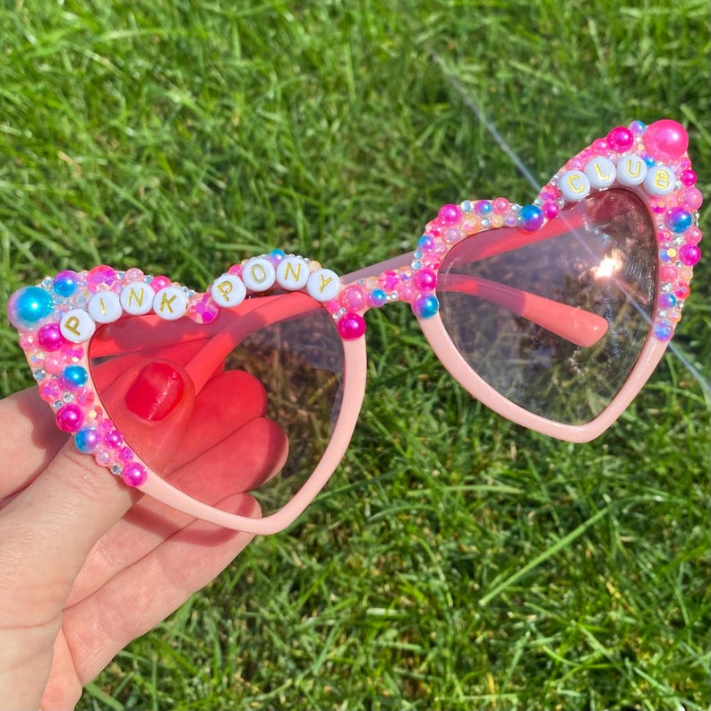 May include: Pink heart-shaped sunglasses with pink, blue, and white beads and the words "Pink Pony Club" spelled out in gold letters.