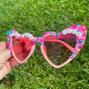 May include: Pink heart-shaped sunglasses with pink, blue, and white beads and the words "PINK PONY CLUB" spelled out in gold letters.