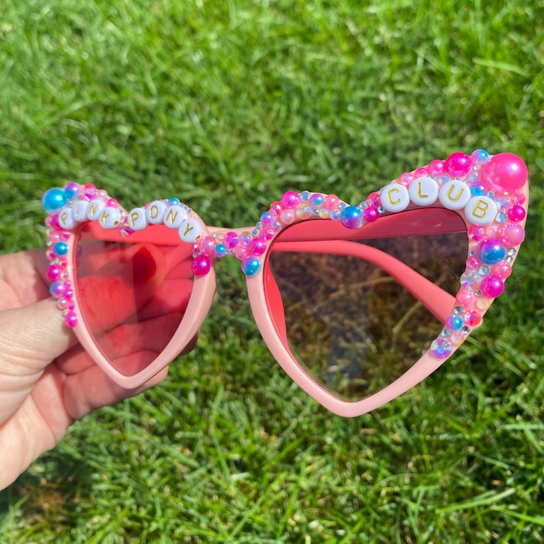 May include: Pink heart-shaped sunglasses with pink, blue, and white beads and the words "PINK PONY CLUB" spelled out in gold letters.