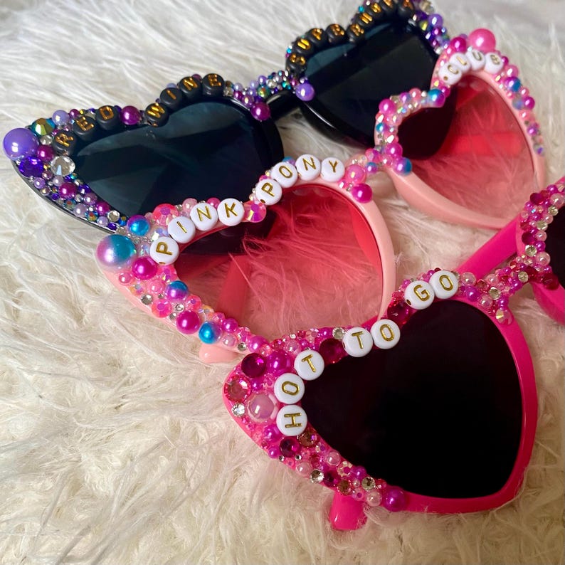 May include: Three pairs of heart-shaped sunglasses with black lenses. The sunglasses are decorated with colorful beads and have words spelled out on the frames, including "Pink Pony", "Hot To Go", and "Freedom Winner".