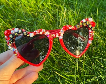 May include: Red heart-shaped sunglasses with black lenses. The frames are embellished with red, white, and gold beads and rhinestones. The word "RED" is spelled out on the left side of the frame, and "ORRES" on the right side.