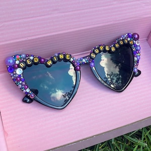 May include: Heart-shaped sunglasses with black frames and lenses. The frames are decorated with purple and gold beads and the words "Red Wine Supernova" are spelled out in gold letters.
