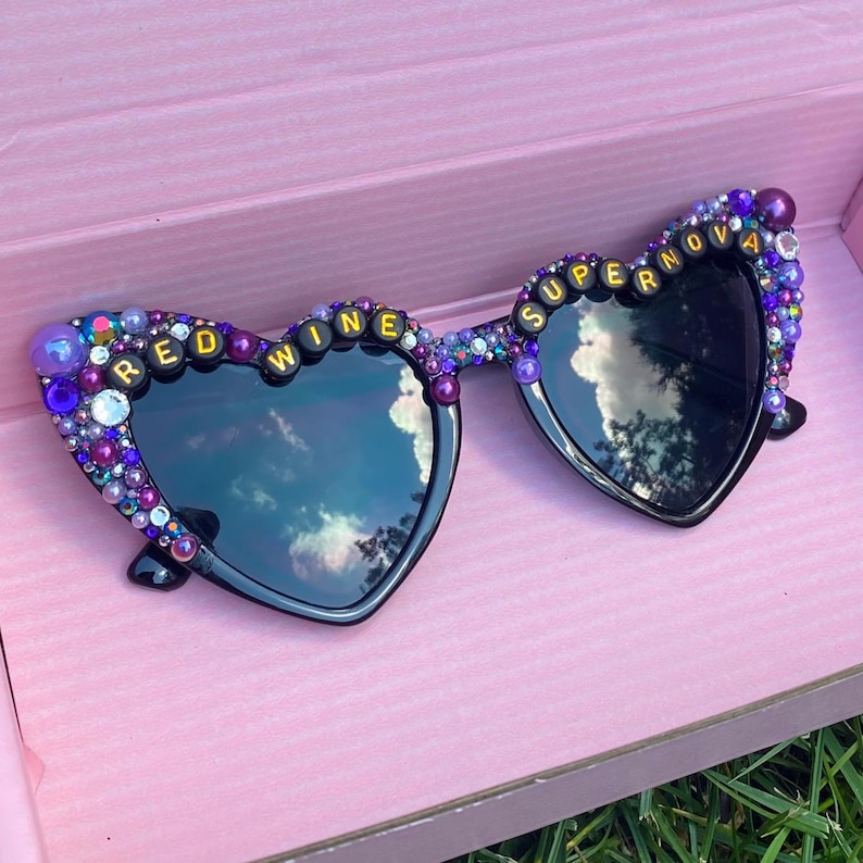 May include: Heart-shaped sunglasses with black frames and lenses. The frames are decorated with purple and gold beads and the words "Red Wine Supernova" are spelled out in gold letters.
