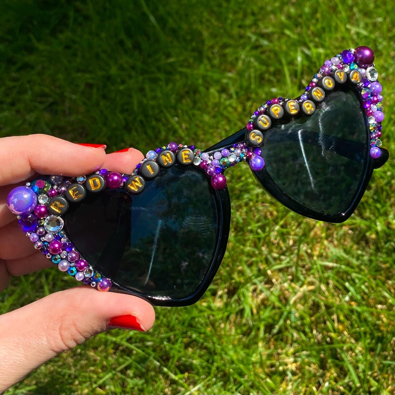 May include: Heart-shaped sunglasses with black frames and lenses, decorated with purple and iridescent beads. The beads spell out "RED WINE IS SUPERIOR" in gold letters.