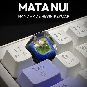 Op de afbeelding: Een close-up van een toetsenbord met een aangepaste hars toetsenkap. De toetsenkap is transparant met een blauw en groen ontwerp. De tekst "MATA NUI HANDMADE RESIN KEYCAP" staat bovenaan. Andere toetsen zijn wit en paars.