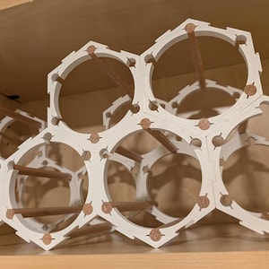 May include: White hexagonal wine rack with wooden dowels. The rack is made of interlocking pieces, creating a honeycomb design. The dowels provide support for the wine bottles. The rack is designed to hold multiple bottles and is made of a sturdy material.
