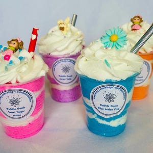 May include: Four colorful bubble bath bombs shaped like ice cream sundaes. Each bomb is in a clear plastic cup, topped with white foam, and decorated with a small figurine and sprinkles. The cups are pink, purple, blue, and orange, with text labels.
