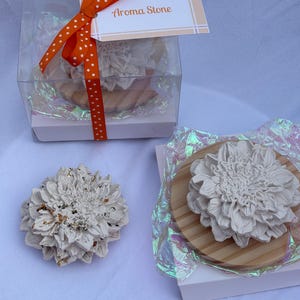 May include: Two white flower-shaped aroma stones, one inside a clear gift box with an orange polka dot ribbon and a tag reading Aroma Stone, and one placed on a wooden base. Both stones feature intricate petal details and iridescent paper.