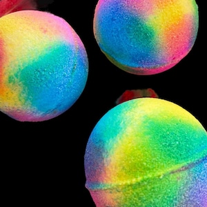May include: Three spherical bath bombs featuring a vibrant, multi-colored tie-dye pattern with swirls of bright yellow, electric blue, magenta, and lime green. Each round bath bomb has a textured, porous surface and a visible seam around the center.