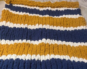 May include: A striped knitted throw blanket in shades of mustard yellow, white, and navy blue. The blanket is made with a chunky yarn and has a soft, fluffy texture.