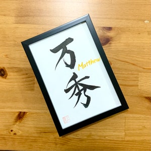 May include: A black framed print with a white background. The print features black calligraphy in Japanese characters and the name "Matthew" written in gold lettering.