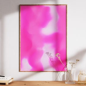 Könnte beinhalten: Ein gerahmter Kunstdruck mit einem lebendigen rosa-weißen abstrakten Design. Der Holzrahmen umgibt das Kunstwerk, das verschwommene Farbverläufe aufweist. Der Druck wird auf einer Holzoberfläche mit dekorativen Elementen präsentiert.