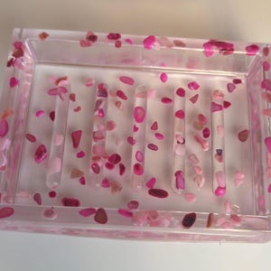 May include: A rectangular soap dish made of clear resin, embedded with pink and magenta stones. The dish has vertical bars and is filled with small, decorative stones in varying shades of pink. The overall design is a blend of functionality and aesthetic appeal.