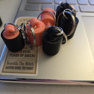 May include: A set of six keychains, three are black and three are pink. The keychains are made of plastic and have a cylindrical shape. The keychains are attached to silver key rings.