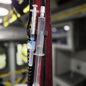 May include: Two clear plastic syringes with blue and white accents are attached to a red leather strap. The syringes are hanging vertically and are partially filled with a clear liquid.