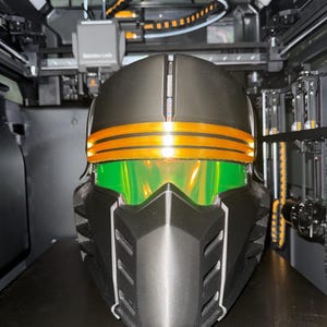 May include: A black and orange helmet with a green visor is displayed inside a 3D printer. The helmet has orange stripes and a detailed, angular design. The printer's interior is visible, with various mechanical components and the text "Bambu Lab".