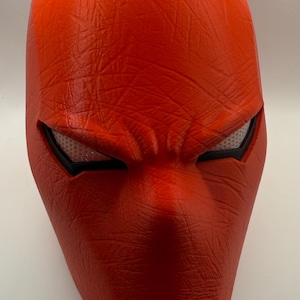 May include: A bright red mask with a textured surface, featuring eye openings with white mesh and black borders. The mask has pointed ears and a prominent nose, resembling a superhero or comic book character.