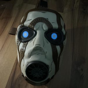 May include: A white and brown mask with glowing blue eyes, resembling a character from a video game. The mask has a gas mask-like design with a black mouth piece and brown accents. The eyes have a mesh design.