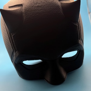 May include: A black mask with pointed ears and white eye openings. The mask has a textured surface and is likely made of a durable material. The design suggests a superhero or cosplay theme.
